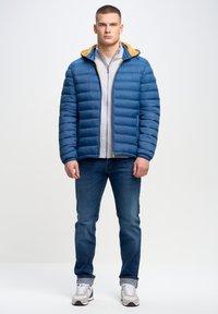 Herre blå pufferjakke med en gul hætte, grå zip-up skjorte under, blå jeans og hvide sneakers med grå detaljer.