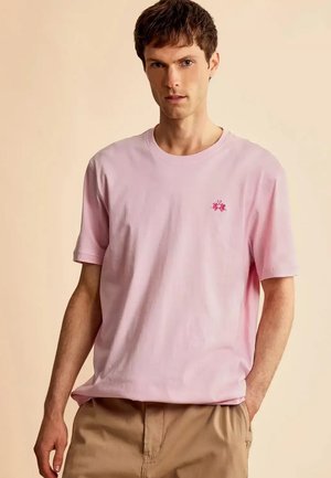 Junger Mann trägt ein hellrosa kurzärmeliges T-Shirt mit kleinem roten, gesticktem Logo auf der Brust, kombiniert mit beigen Hosen, steht vor neutralem Hintergrund.