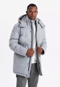 Chaqueta acolchada gris con capucha, que cuenta con un cierre de cremallera y a presión, bolsillos laterales y una textura suave, usada sobre una camisa blanca.