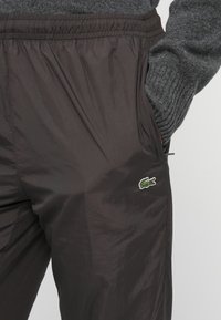 Person trägt dunkelbraune Hose mit elastischem Bund und grünem Krokodil-Logo, Hand in der Tasche, kombiniert mit grauem Strickpullover.