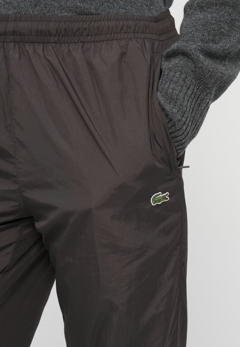 Person med mörkbruna byxor med elastisk midja och grön krokodil-logotyp, handen i fickan, kombinerat med en grå stickad tröja.