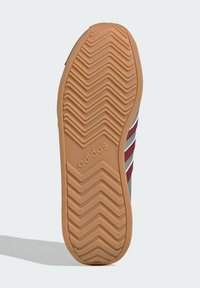 Rubber sneaker sole featuring a zigzag tread pattern, tan color with a logo, bordered by a grey upper with red and white stripes.