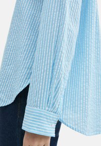 Camicia azzurra chiara con righe verticali bianche, caratterizzata da un orlo curvo e dettagli sui polsini. Realizzata in tessuto testurizzato.