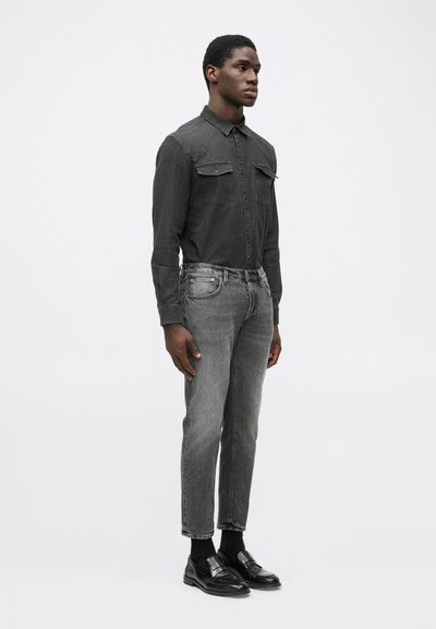 Camicia di denim grigia con due tasche sul petto, abbinata a jeans grigi a gamba affusolata e mocassini neri. Il modello è in piedi contro uno sfondo semplice.