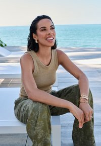 Débardeur en bronze côtelé, pantalon à jambes larges à motifs vert olive, boucles d'oreilles et bracelets dorés, assise devant un paysage en bord de mer.