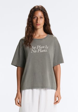 Kobieta z długimi falującymi włosami, ubrana w luźny szary T-shirt z tekstem "The Plan Is No Plans" oraz białe spodnie, stojąca na jednolitym tle.