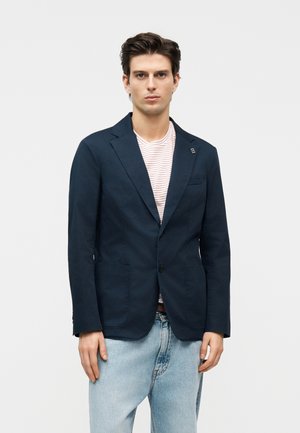 Ung mand iført en navy blazer over en hvid og rødstribet T-shirt kombineret med lyseblå jeans, stående foran en ensfarvet baggrund.