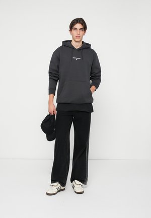 Tmavě šedá mikina s kapucí a přední kapsou, doplněná širokými černými kalhotami. Bílé tenisky s černými pruhy dokončují tento outfit.