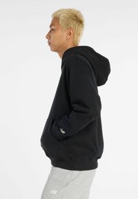 Sudadera negra con bolsillo canguro, puños y dobladillo acanalados. Presenta un detalle de logo en la manga. La persona lleva pantalones de chándal gris claro.