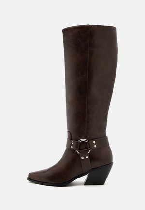 Botte haute en cuir marron foncé avec un bout pointu, un talon bas incliné et une sangle décorative avec anneau en argent et rivets autour de la cheville.