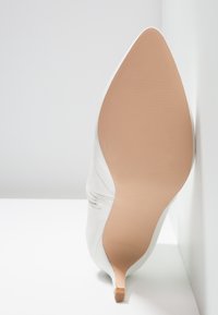 Chaussure à talon haut blanche à bout pointu montrée semelle vers le haut, révélant une semelle beige lisse et un talon aigu fin sur fond blanc.