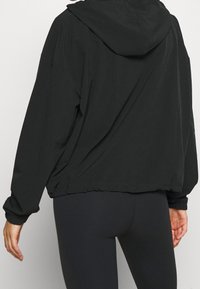 Sort hættejakke med løs pasform, indsnøring i kanten og elastiske manchetter. Glat, letvægtsstof uden synligt hardware eller mønstre.