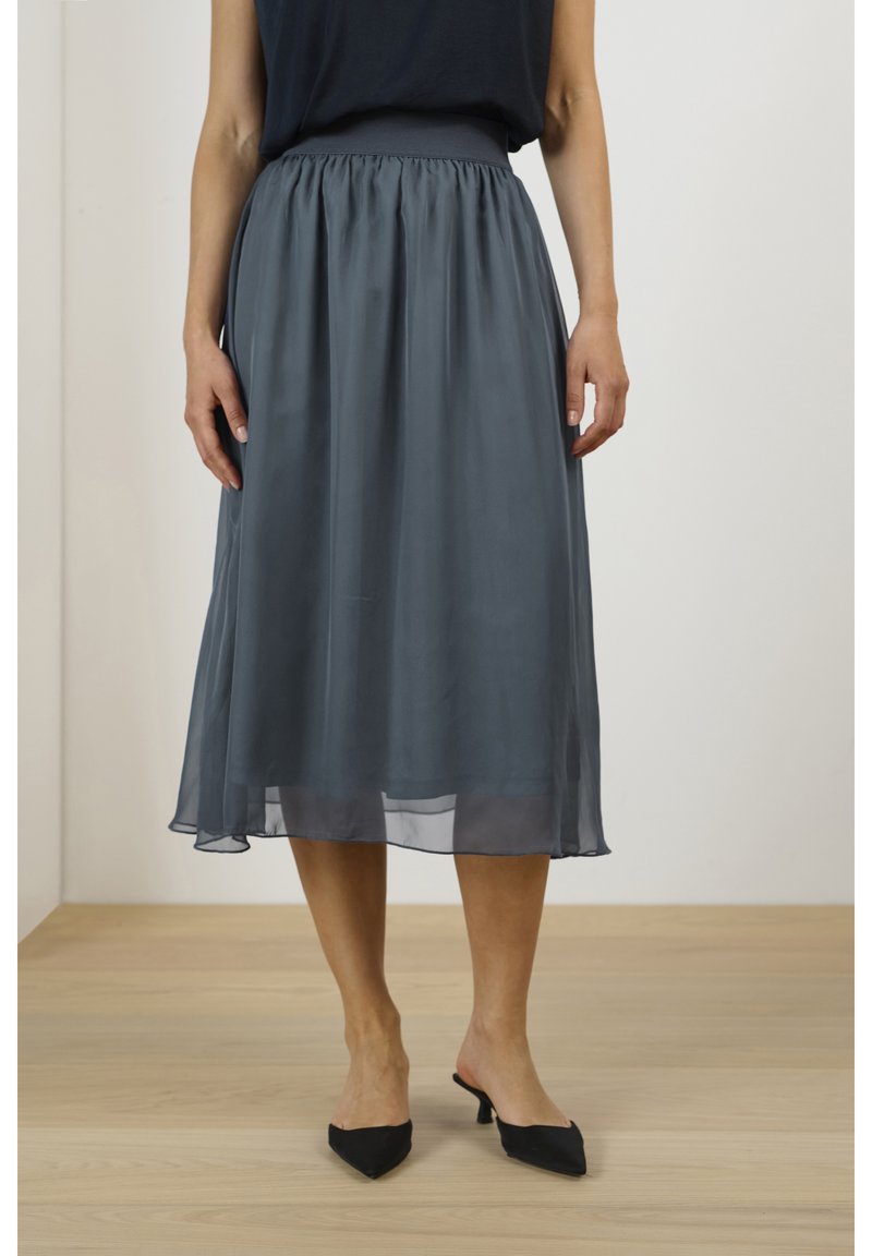 Femme portant une jupe transparente gris foncé jusqu'aux genoux avec des escarpins noirs à bout pointu, debout sur un sol en bois.