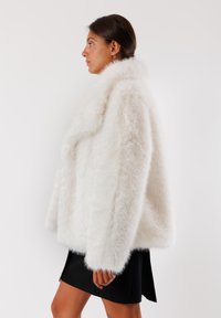 Cappotto in pelliccia sintetica bianca con ampio colletto, vestibilità oversize e maniche lunghe. Texture morbida e aspetto voluminoso, abbinato a una gonna nera.