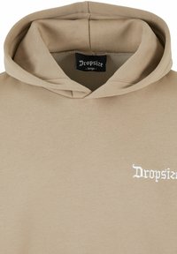 Sudadera con capucha beige, hecha de un material suave. Presenta un bolsillo delantero, cordones y un logo bordado en blanco que dice "Dropsize" en el pecho.