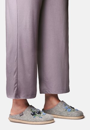 Pieds portant des chaussons en laine grise avec une broderie florale bleue et noire, associés à un pantalon lavande ample sur un fond uni.