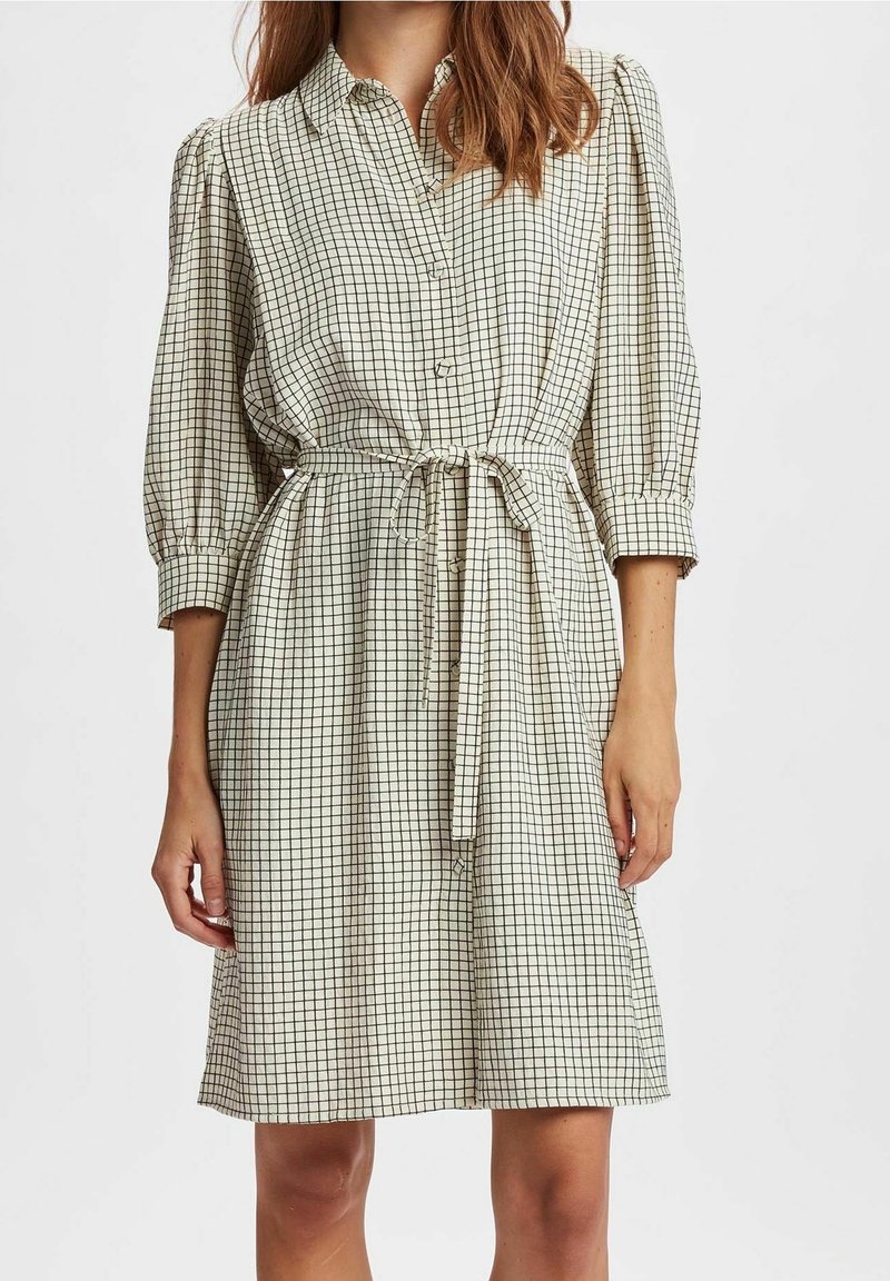 Vestito a scacchi in crema e nero, con chiusura frontale a bottoni, vita legata e maniche a palloncino. Realizzato in tessuto leggero con una vestibilità comoda.