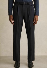Mann in maßgeschneiderten schwarzen Anzughosen mit Bundfalten, einem Gürtel und schwarzen Leder-Brogues, der vor einem neutralen Hintergrund steht.