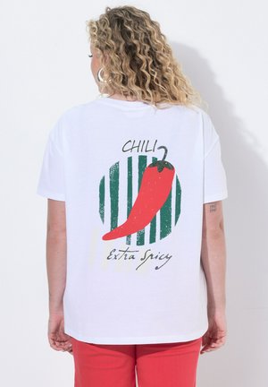 Persoon met krullend haar die een wit T-shirt draagt met een rood chili-beeld en tekst "CHILI Extra Spicy" op de achterkant, gecombineerd met een rode broek.