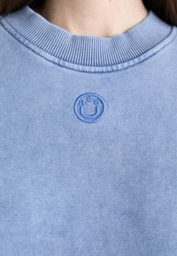 Gros plan sur un sweat-shirt à col rond côtelé bleu clair avec un petit visage souriant brodé et les lettres « H G C » sur la poitrine.