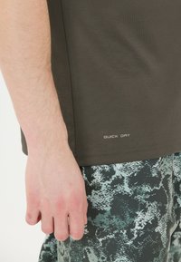 Lähedalt vaadatuna on käsi tumeda oliivroheline kiire kuivatamise särgi kõrval ning roheliste ja hallide abstraktse mustriga pükste kõrval.