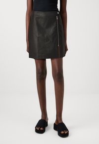 Mini-jupe en cuir noir à texture lisse, avec une fermeture éclair latérale et un ourlet net. Associée à des sandales plates noires.