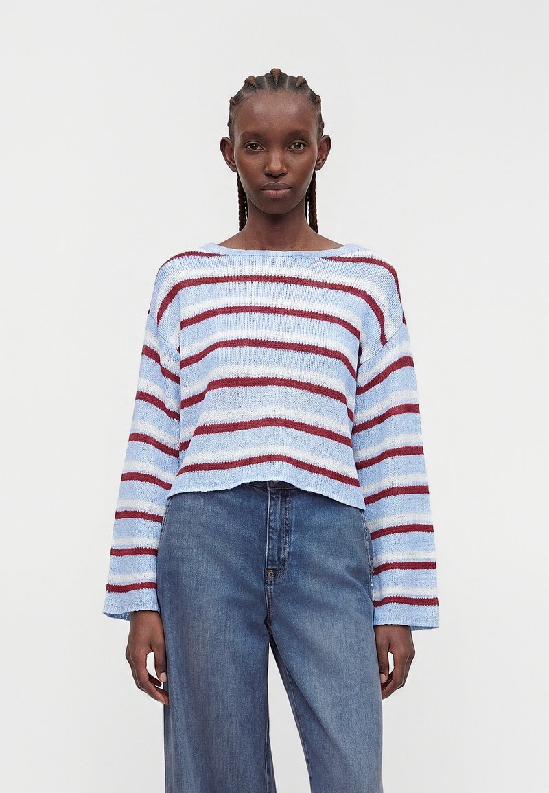 Femme aux cheveux tressés portant un pull rayé bleu clair et bordeaux et un jean bleu taille haute sur un fond uni.