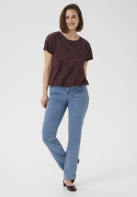 Kurzarmoberteil mit einem dunklen burgunderfarbenen Blumenmuster, kombiniert mit hellblauen Jeans im geraden Schnitt. Das Model steht vor einem neutralen Hintergrund.