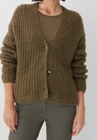 Olivgrön stickad cardigan med kraftig textur, djup V-ringning och tre svarta knappar fram. Långa, lätt puffade ärmar.