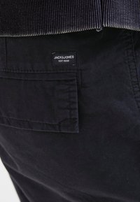 Černé bavlněné kalhoty s boční kapsou a přišitou nálepkou s nápisem "JACK & JONES EST.1990." Mají hladkou texturu a odolnou tkaninu.