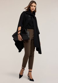 Cappotto nero con colletto oversize, abbinato a una blusa nera a trama, pantaloni marroni aderenti e décolleté neri a punta. Tiene in mano una borsa nera.