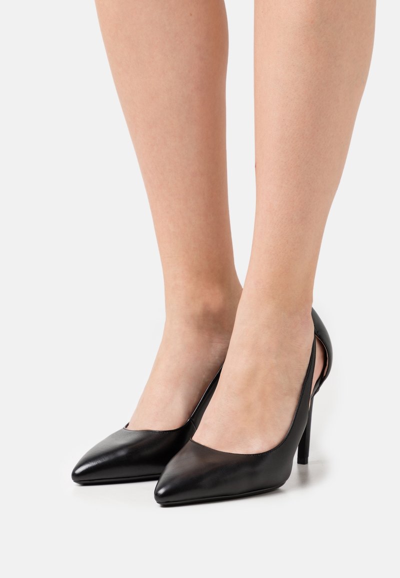 Escarpins noirs à bout pointu portés pieds nus, montrant les jambes et les chevilles sur un fond clair uni.