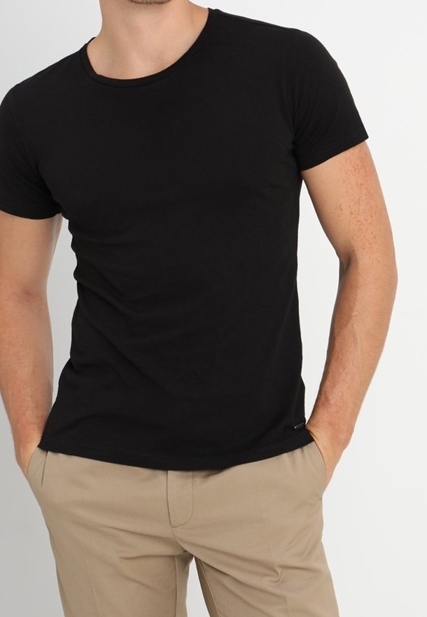 Homme portant un t-shirt noir ajusté à manches courtes et un pantalon beige, debout les mains dans les poches devant un fond blanc uni.