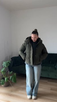 Mladá žena v zelenej nadýchanej bunde a voľných džínsoch stojí v interiéri pri zelenej pohovke a kvetináči na drevenej podlahe.