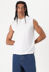 T-shirt bianca senza maniche in cotone con scollatura rotonda e un piccolo logo sul petto. Abbinata a jeans in denim blu. Design semplice e casual.