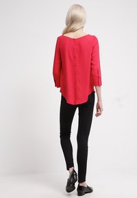 Blusa roja de mangas tres cuartos, escote redondo y cierre de botón en la parte posterior. Combinada con jeans ajustados negros y zapatos brillantes.
