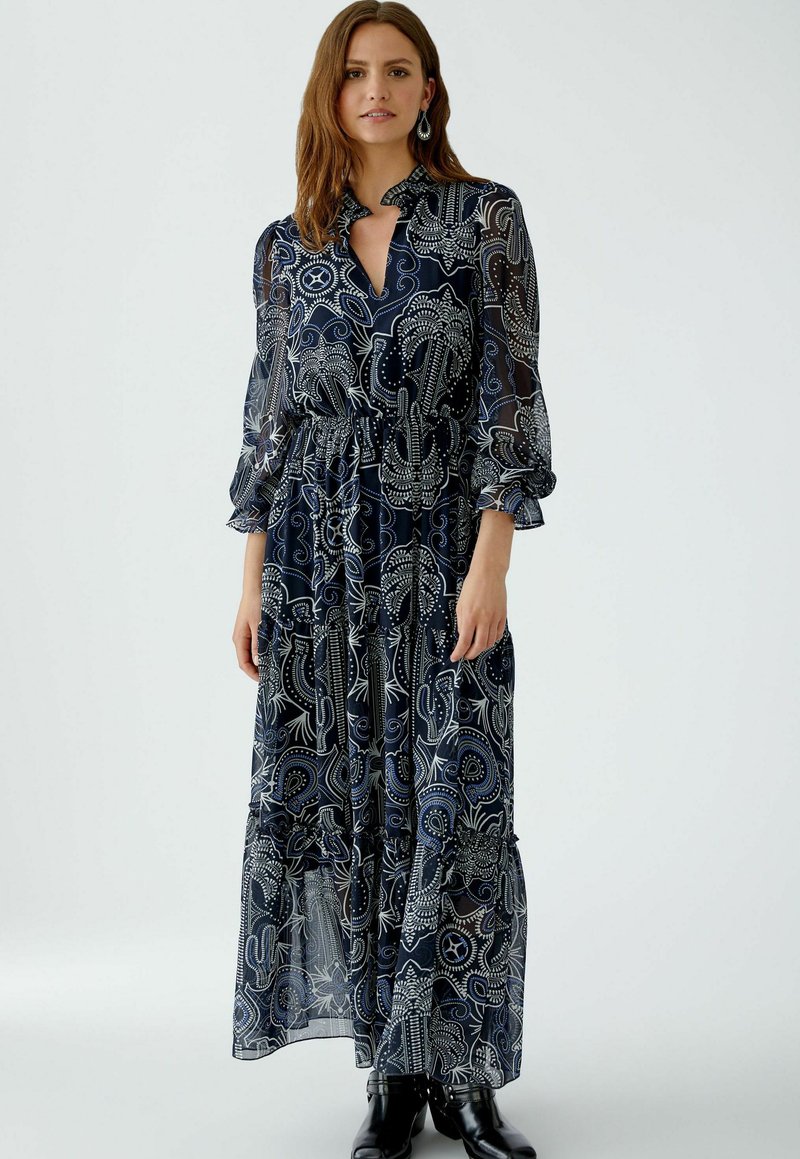 Langes, dunkelblaues Kleid mit einem Paisleymuster in Weiß und Hellblau. Transparente Ärmel mit Rüschenausschnitten und einer taillierten Taille. Hoher Halsausschnitt.