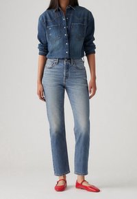 Camisa de mezclilla oscura con cierre de botones, dos bolsillos frontales, combinada con pantalones vaqueros de pierna recta en azul claro. Completado con zapatos planos rojos.