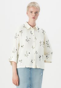 Vrouw met kort blond haar die een gebroken wit overhemd met zwarte bloemenprint en een lichtblauwe denim rok draagt.