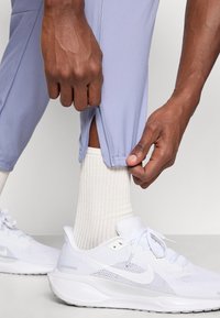 Χέρια ρυθμίζουν τον φερμουάρ στον αστράγαλο ενός μωβ αθλητικού παντελονιού, το οποίο φοριέται με λευκές ριγέ κάλτσες και λευκά αθλητικά παπούτσια Nike.