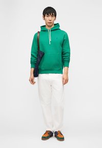 Mlajši moški, oblečen v zelen pulover s kapuco, bele hlače, večbarvne čevlje in z mornarsko modro torbo čez eno ramo.