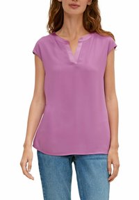 Kobieta w krótkim rękawie lawendowej bluzki z dekoltem w serek, w połączeniu z niebieskimi dżinsami, pokazana od ramion do bioder.