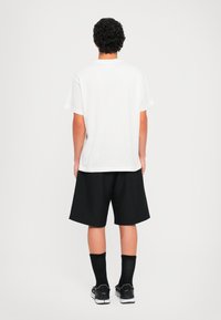 Pessoa com cabelo encaracolado está de costas, vestindo uma t-shirt branca simples, calções pretos, meias pretas e ténis pretos.