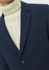 Marinblå blazer med ett notchsnyggt slag och en enkel svart knapp. Under finns en krämfärgad ribbad polotröja med ett kabelstickat mönster.