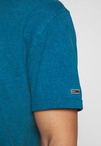 Camiseta de algodón de color verde azulado de manga corta con cuello redondo, que presenta un pequeño parche de logo en la manga y detalles de costura visibles.