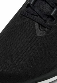 Zapatilla deportiva negra con parte superior de punto texturizado, cordones planos y suela acolchada, con patrones acanalados distintivos y acentos sintéticos.