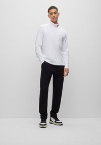 Camisa blanca de manga larga con cuello alto, combinada con pantalones negros y zapatillas multicolor con acentos en rojo y gris.