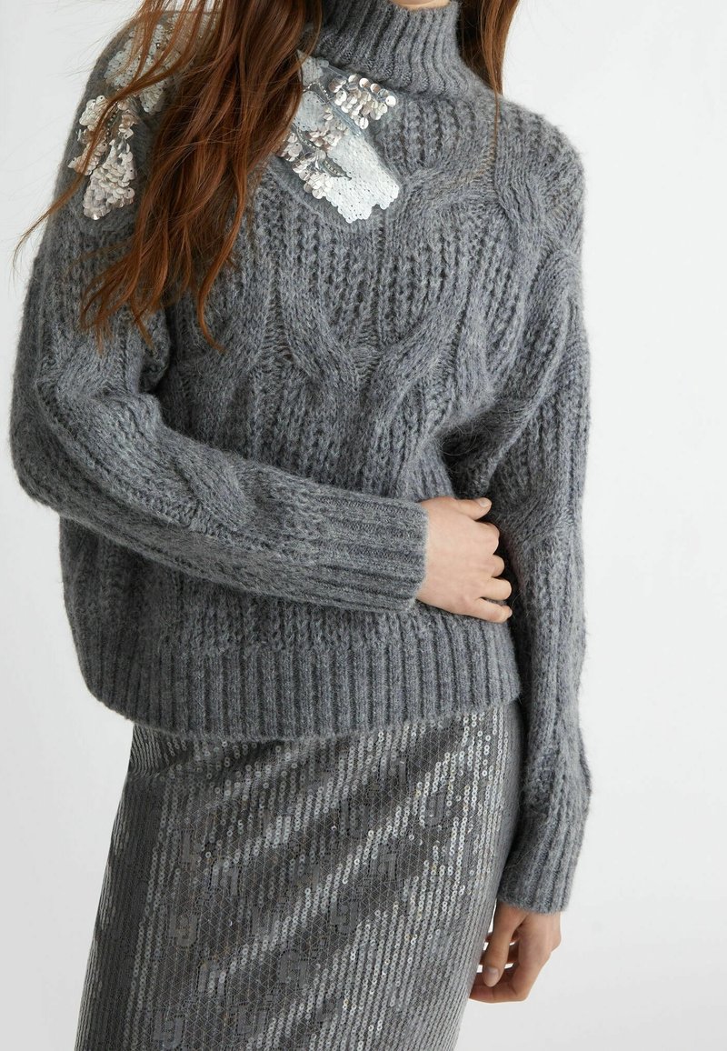 Grau gestrickter Pullover mit einem gerippten Saum und Bündchen, der ein pailettenbesetztes Detail an der linken Schulter hat. Die Textur ist weich und kuschelig.