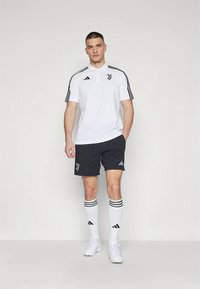 Λευκό πόλο πουκάμισο με μαύρες ρίγες και λογότυπο, σκούρα σορτς με λογότυπο, λευκές κάλτσες με μαύρες ρίγες και λευκά αθλητικά παπούτσια.