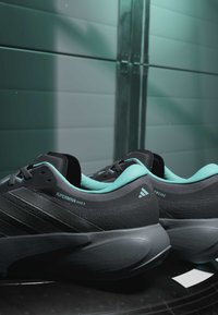 Paire de chaussures de course Adidas Supernova Rise 3 noires et bleu sarcelle placées côte à côte sur une surface réfléchissante devant un mur carrelé gris.